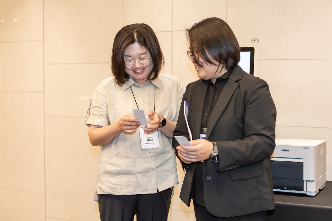 즉석 인화된 사진을 함께 확인하는 참석자 두 명, 옆에 프린터가 보임